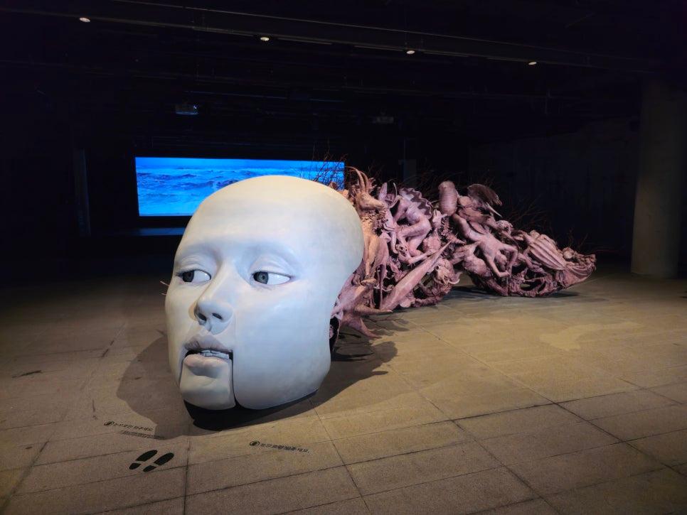 흰색 얼굴 조형물과 분홍색 인체 조각이 배치된 전시 공간