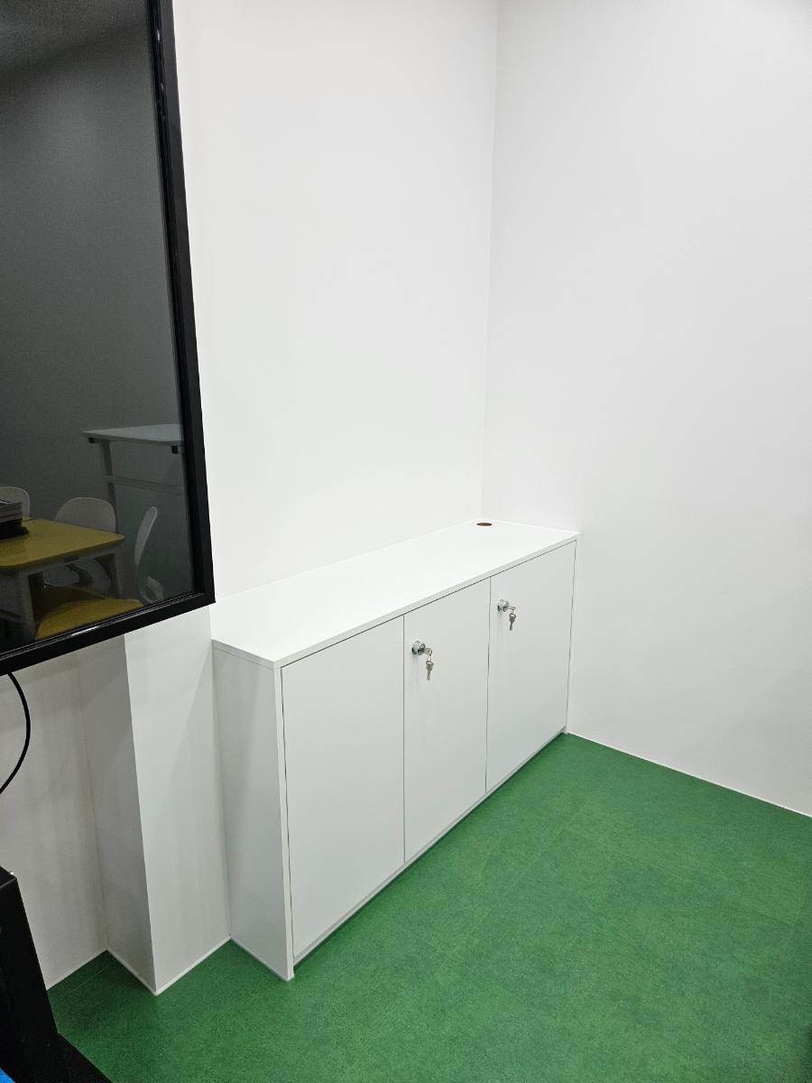 흰 벽, 녹색 바닥, 흰색 수납장이 있는 실내 공간. 왼쪽에는 검은 테두리의 유리문이 보이며, 유리문을 통해 다른 공간의 노란색 테이블과 흰색 의자가 보인다.