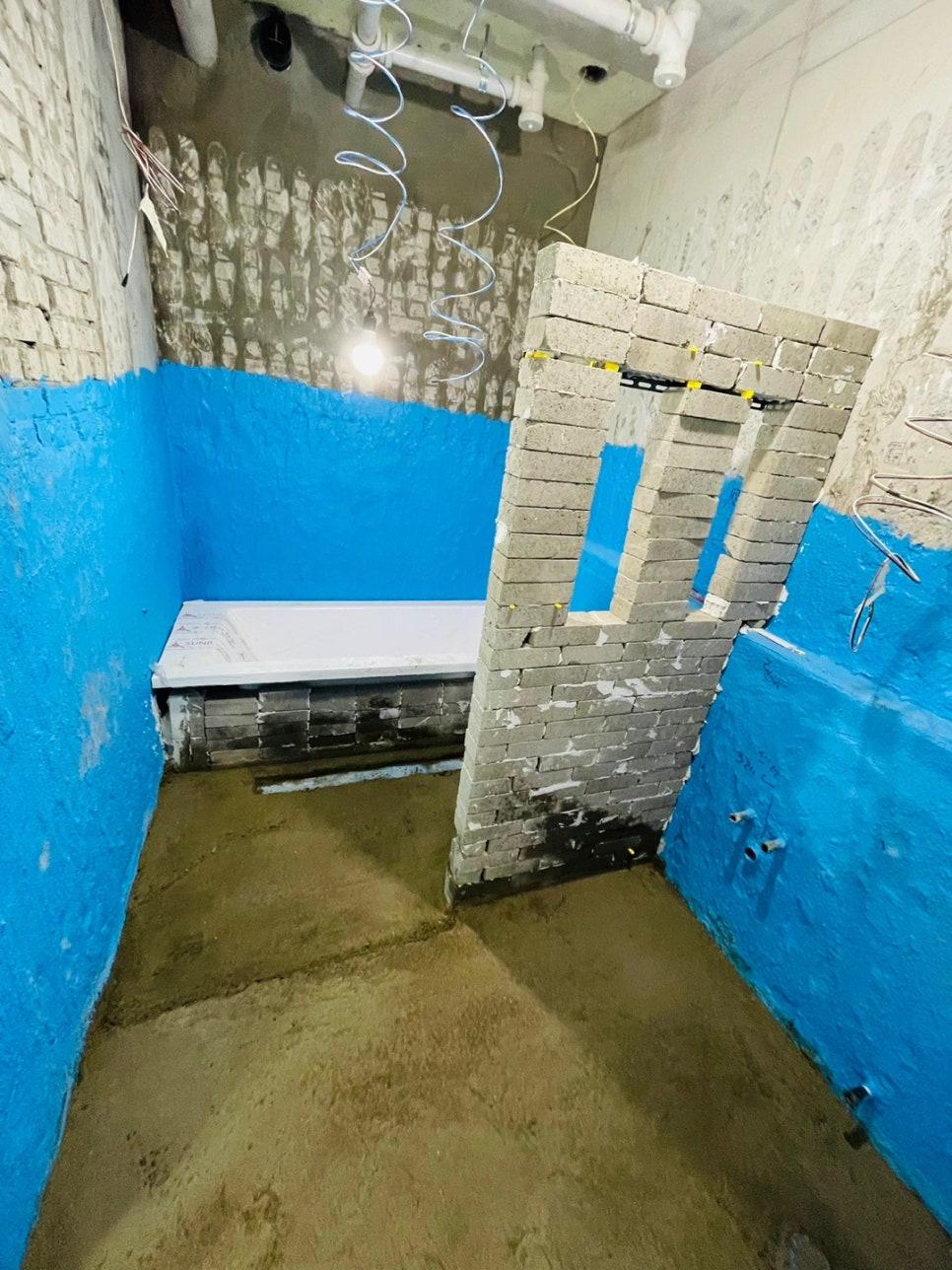 실내 공간에 파란색 벽면, 벽돌 구조물, 흙바닥, 천장의 배관 및 전선이 보이는 공사 중인 공간