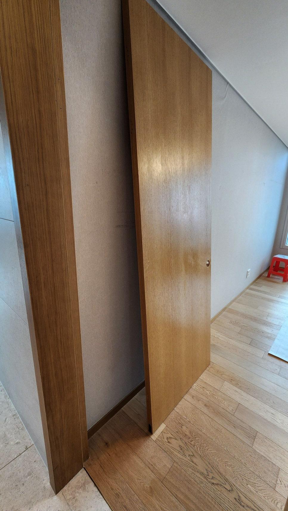 복도 공간에 목재 마감의 문이 열린 상태로 보이며, 바닥은 목재 바닥재, 벽은 벽지로 마감된 공간