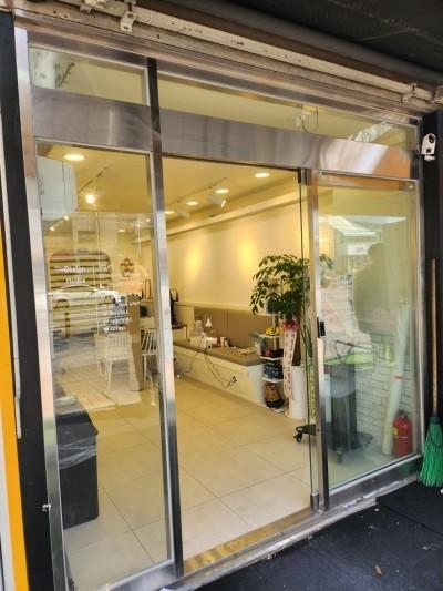 실내 공간의 유리문과 내부를 보여주는 사진. 내부에는 타일 바닥, 벽면 조명, 식물, 수납장, 의자, 쓰레기통, 화재 진화기 등이 보임. 유리문은 슬라이딩 구조로, 문 위쪽에는 금속 프레임이 보임.