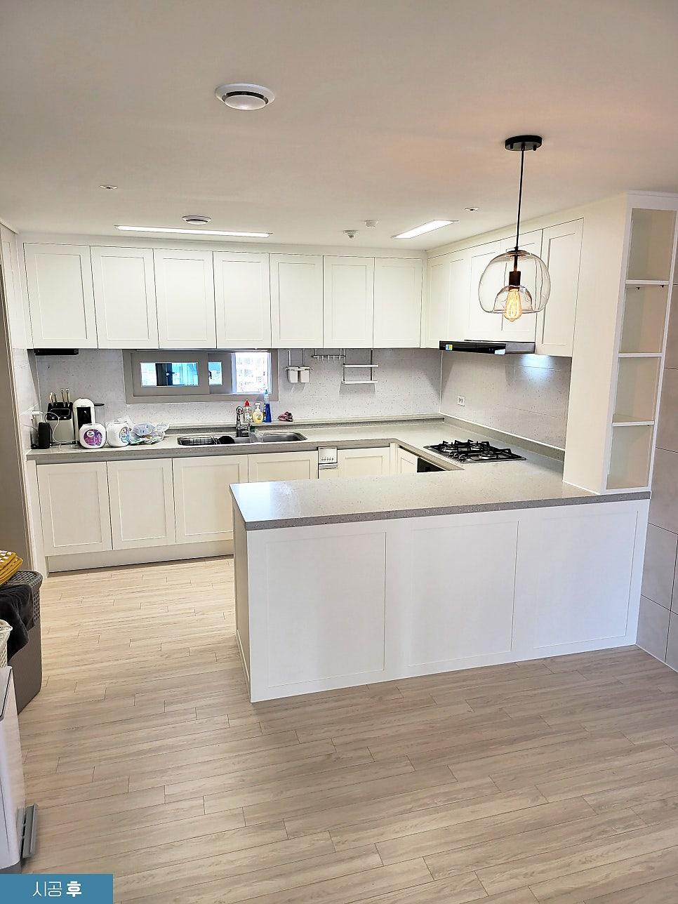 흰색 상부장과 하부장, 중앙 아일랜드 카운터, 스테인리스 싱크대, 가스레인지, 벽면 타일, 목재 바닥재, 천장 조명, 유리 램프가 보이는 주방 공간