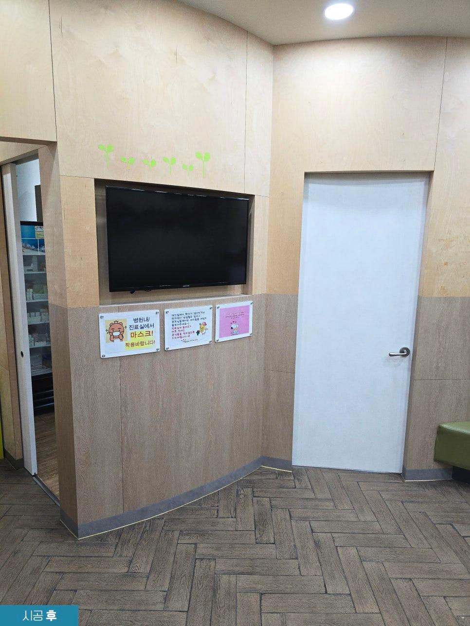 실내 벽면에 TV 설치, 하단에 3장의 안내문구가 부착된 공간. 벽은 목재 패널로 마감, 바닥은 나무 패턴 타일. 우측에 흰색 도어, 좌측에 문이 열린 공간 보임.
