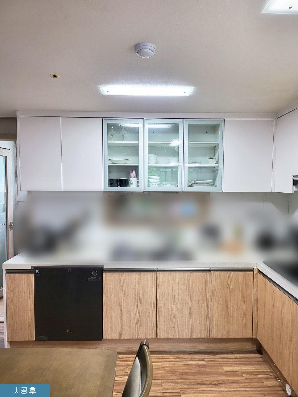 하얀색 상부 수납장과 나무 질감 하부 수납장이 있는 주방. 상부 수납장 중 3개는 유리 문으로 되어 있으며, 내부에 접시 등이 보입니다. 하부 수납장 중 왼쪽에는 검은색 세탁기 또는 건조기, 오른쪽에는 싱크대가 보입니다. 바닥은 나무 패턴의 바닥재로 되어 있으며, 천장에는 조명과 소방 장치가 있습니다. 왼쪽에는 문이 보입니다.