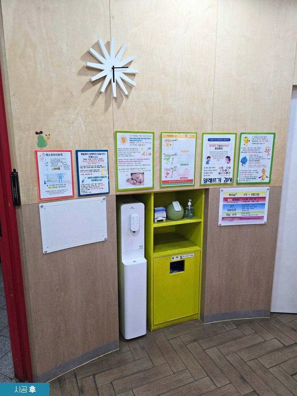 벽면에 흰색 별 모양 시계, 여러 종류의 안내문과 포스터가 부착된 공간. 중앙에 흰색 물 dispensers, 노란색 수납장과 쓰레기통이 설치되어 있으며, 바닥은 나무 패턴의 타일로 마감됨.