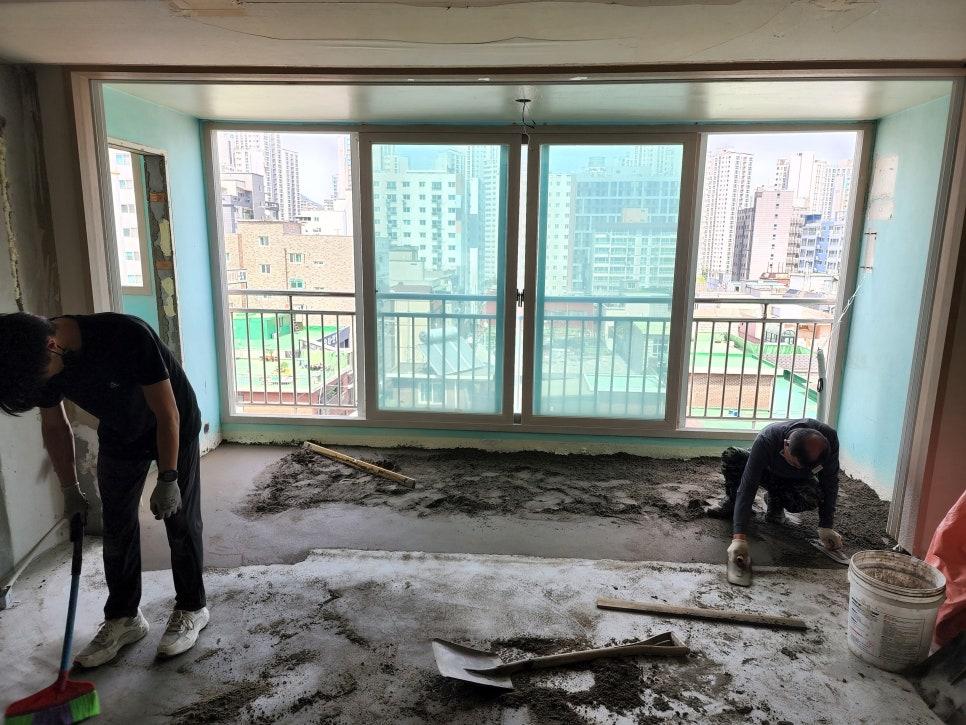 실내 공사 중인 공간, 벽은 파란색 도배, 바닥은 흙과 자재가 흩어져 있고, 창문 근처에 작업 중인 노동자 2명, 창문 외부에는 고층 건물이 보임
