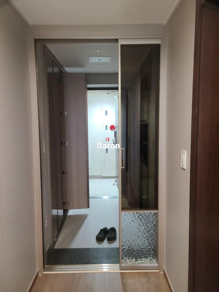 현관 공간에 슬라이딩 도어가 설치되어 있으며, 도어 왼쪽에는 수납장이 있고, 오른쪽에는 유리 패널과 투명 유리 패널이 있는 도어가 있습니다. 도어 내부에는 검은색 슬리퍼가 놓여 있고, 바닥은 목재 마루와 타일로 구성되어 있습니다. 벽은 베이지색 벽지로 마감되어 있으며, 벽에 전등 스위치가 있습니다.