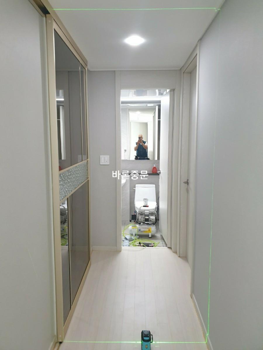 복도 공간, 벽은 흰색, 바닥은 밝은 색 바닥재, 천장에 조명이 설치되어 있으며 좌측에는 유리 문이 있는 수납장이 보임