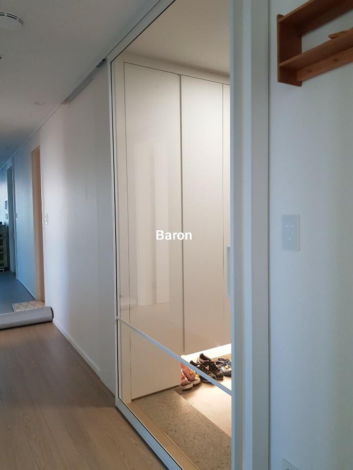 흰 벽, 나무 바닥, 유리 문으로 된 수납장이 있는 복도. 문 안쪽에 신발이 놓여 있고, 멀리서 복도 끝에 다른 방이 보임.