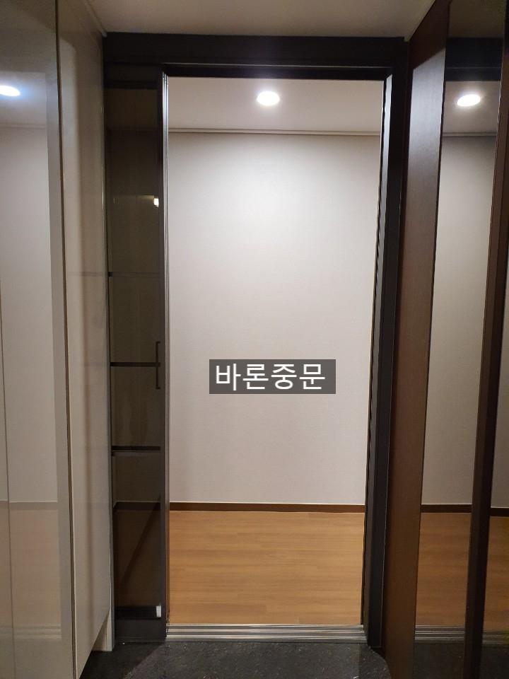 복도 공간, 벽은 상단 흰색 도배, 하단 목재 패널 마감, 천장에 라운드 형광등 조명, 좌측에 유리 문이 있는 수납장, 우측에 문이 있는 공간