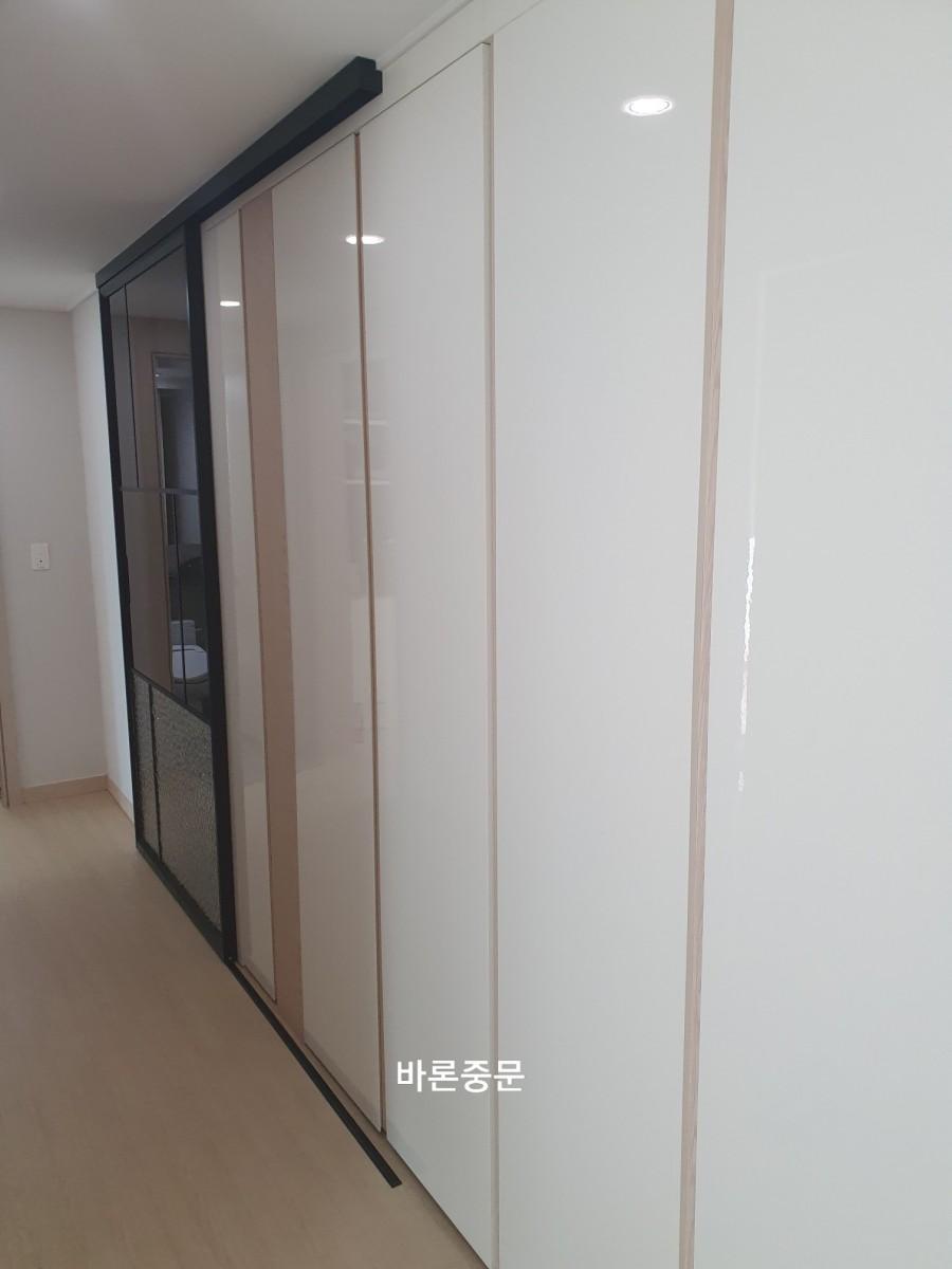 복도 공간에 흰색과 베이지색의 수납장이 설치되어 있으며, 일부 문은 유리 패널로 되어 있고, 바닥은 목재 마루재로 되어 있습니다. 천장에는 내장 조명이 설치되어 있습니다.