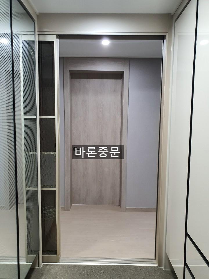 복도 공간에 벽면과 바닥재가 보이며, 좌측에는 유리와 수납장이 있는 문이 열려 있고, 중앙에는 문이 닫혀 있는 공간이 보임