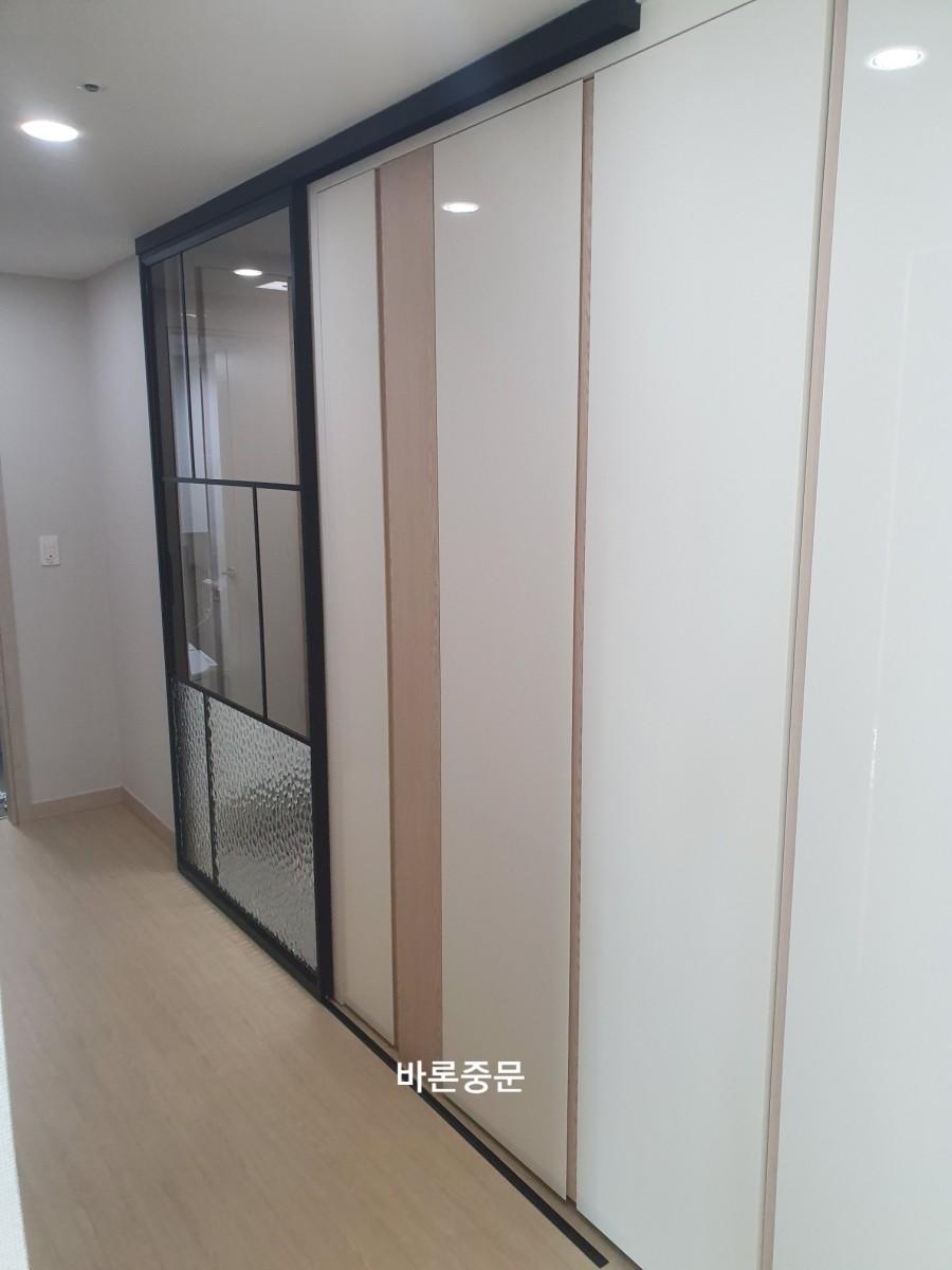 복도 공간에 흰색과 베이지색의 수납장이 설치되어 있으며, 일부 문은 유리 패널로 되어 있고, 바닥은 목재 마감재로 되어 있습니다. 천장에는 LED 조명이 설치되어 있습니다.