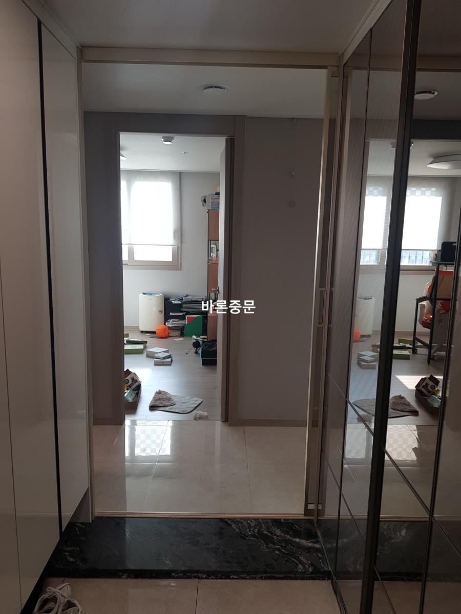 복도 공간에 흰색 벽과 흰색 타일 바닥이 보이며, 양쪽에 유리문과 벽면 수납장이 설치되어 있고, 뒤쪽으로는 거실 공간이 연결되어 있으며, 바닥에 물건들이 놓여 있는 상태
