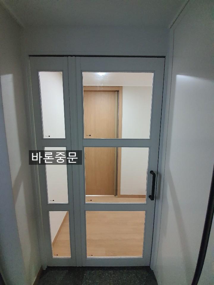 회색 도어가 있는 현관 공간. 도어는 상하로 4개의 유리 패널로 구성되어 있으며, 중앙 유리 패널을 통해 내부의 나무 문이 보임. 벽은 흰색으로 마감되어 있고, 바닥은 어두운 색의 타일로 되어 있음.