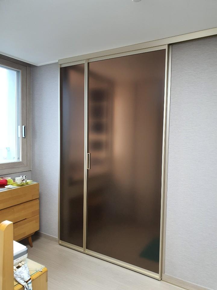 실내 벽지가 보이며, 우측에 슬라이딩 도어가 설치된 공간. 좌측에는 창호가 있고, 창호 아래에는 목재 수납장이 배치되어 있으며, 수납장 위에는 컵과 빨간 물건이 놓여 있다. 바닥은 목재 마루로 마감되어 있다.
