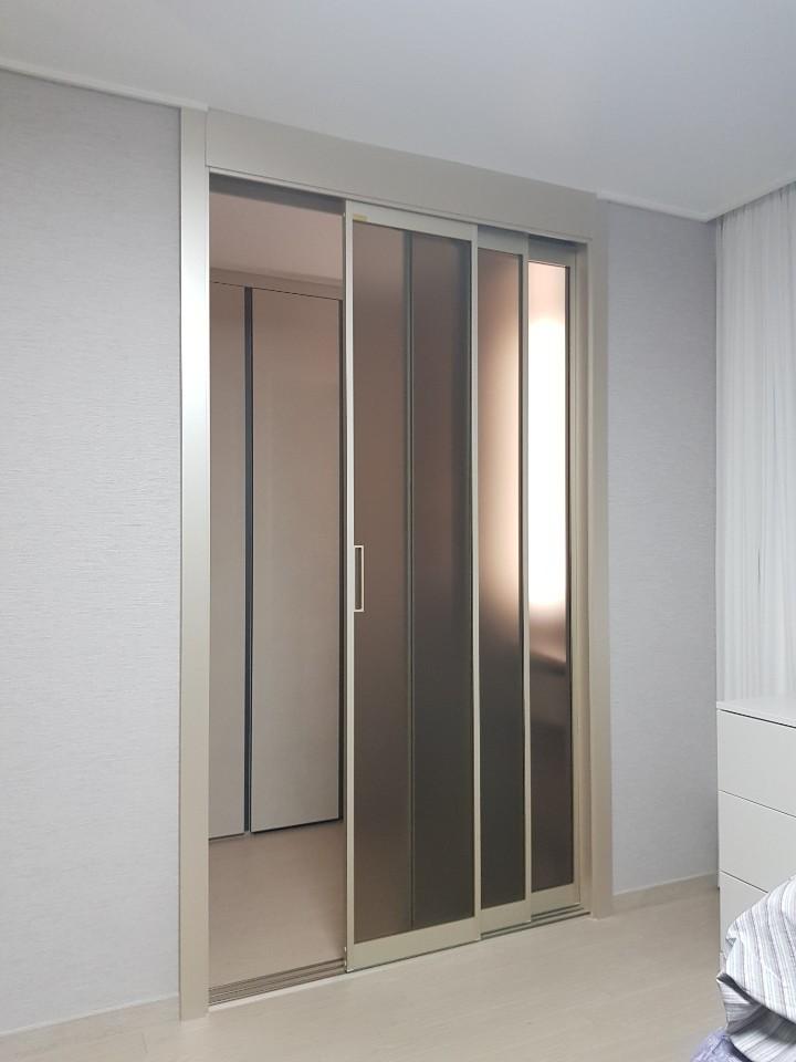 실내 벽면은 흰색 벽지로 마감되어 있으며, 중앙에 슬라이딩 도어가 설치되어 있고, 도어는 반투명 유리 패널로 구성되어 있습니다. 도어 왼쪽에는 다른 방이 보이며, 오른쪽에는 창문이 있는 공간이 보입니다. 바닥은 밝은 색의 마루재로 마감되어 있습니다.