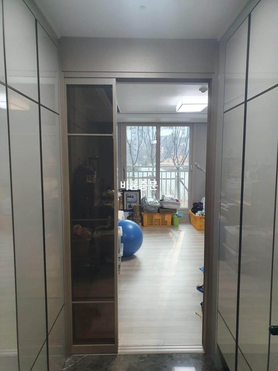 복도 공간에 좌우로 유리문이 있는 수납장이 설치되어 있으며, 뒤쪽에는 창문이 있는 방이 보임. 방 안에는 바구니와 파란색 공, 쓰레기통 등이 놓여 있고, 바닥은 목재재로 마감됨.