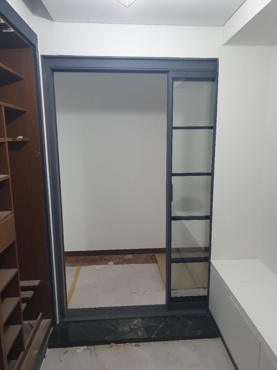 실내 공간에 검은색 프레임의 슬라이딩 도어가 설치되어 있으며, 왼쪽에는 나무 재질의 수납장, 오른쪽에는 흰색 수납장이 보이고, 바닥은 흰색과 검은색의 타일로 마감되어 있으며, 도어 내부 공간은 흰색 벽과 일부 노출된 바닥재가 보입니다.