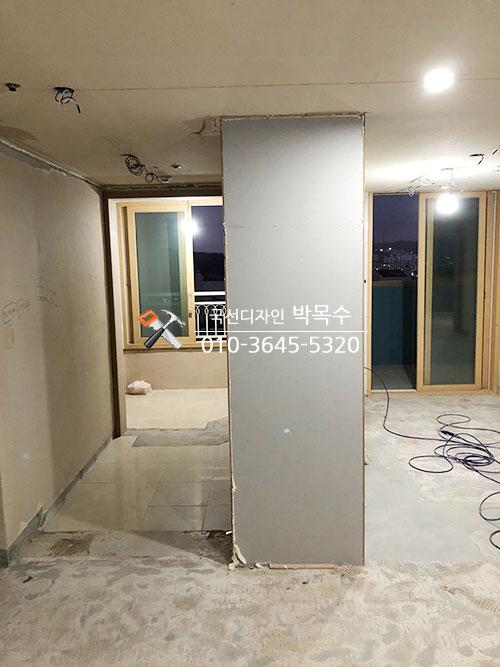 복도 공간, 벽면은 흰색 벽지가 부분적으로 도배되지 않은 상태, 바닥은 타일과 콘크리트가 혼합된 상태, 천장에 전선이 노출되어 있고 중앙에 큰 벽체가 설치 중인 공간