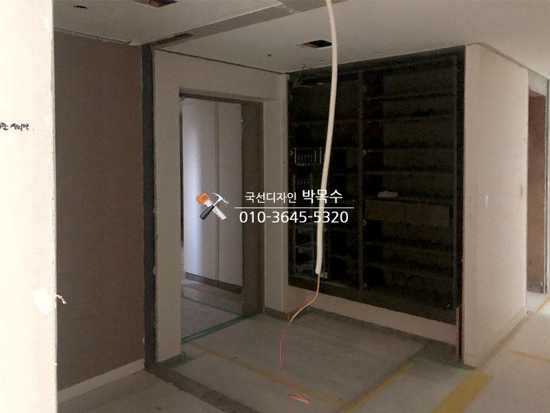 복도 공간에 벽면이 마감되지 않은 상태로, 일부 벽면에 흰색 페인트가 칠해져 있고, 중앙에 유리문이 있는 수납장이 설치되어 있으며, 바닥에는 노란색과 녹색의 테이프가 붙어 있는 공사 중인 공간
