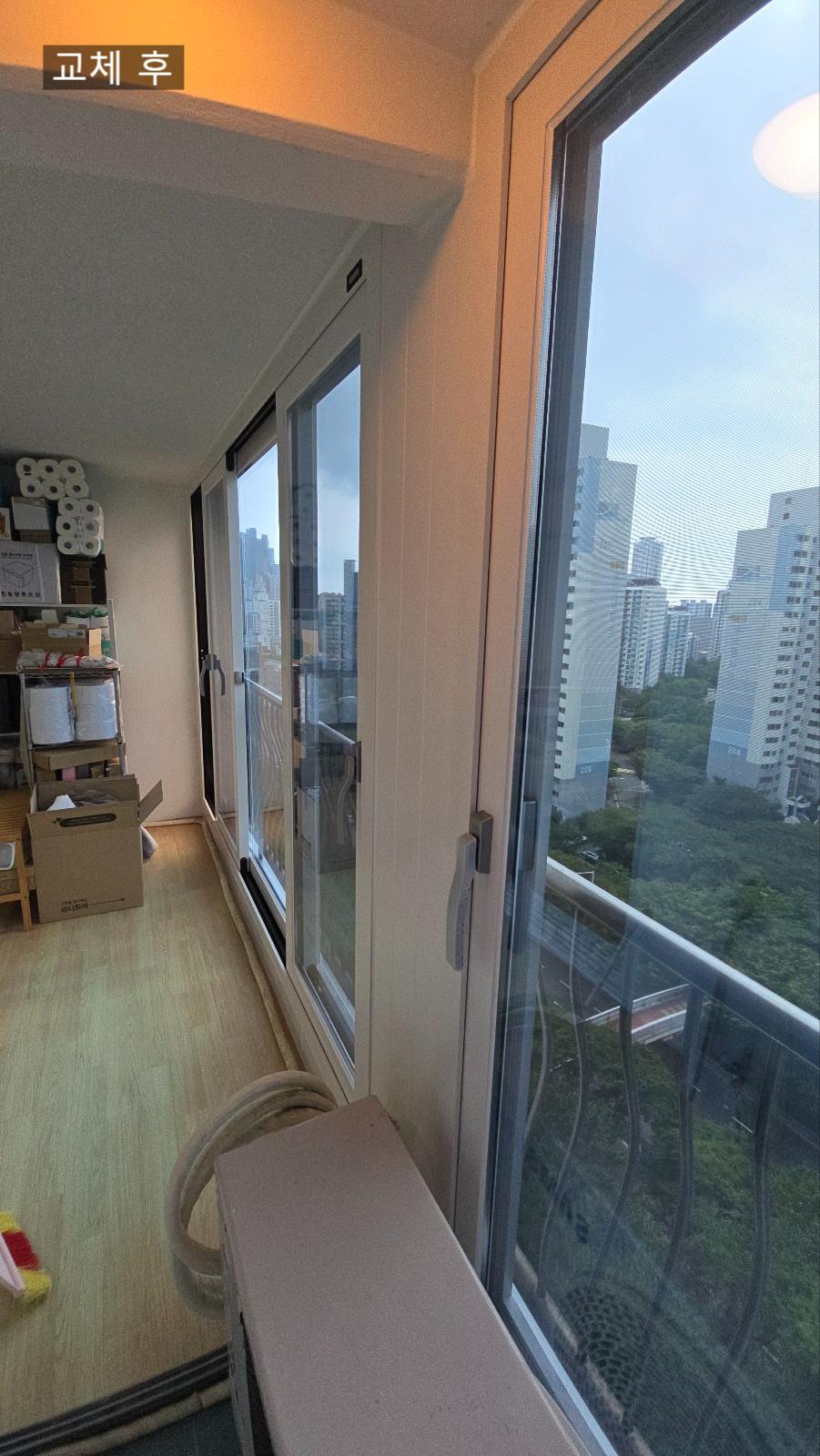 발코니 공간, 벽면에 흰색 도어와 유리창이 설치되어 있으며, 바닥은 목재 마루재로 되어 있고, 왼쪽에는 상자와 롤러 등이 놓인 선반과 상자, 오른쪽에는 유리창으로 된 발코니 문이 보임
