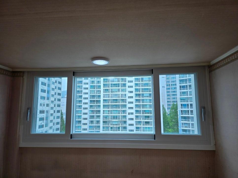 실내 벽면과 천장, 창호가 보이는 창호 근접 촬영. 벽면은 베이지색 벽지, 천장은 베이지색 마감, 창호는 세 개의 유리 패널로 구성된 창문. 창문 외부에는 고층 아파트 건물이 보임.