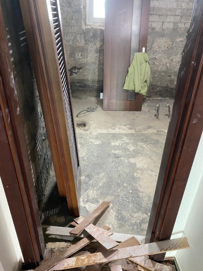 공사 중인 공간으로, 벽은 돌로 된 구조로 마감되지 않았으며, 바닥은 흙과 먼지가 쌓인 상태. 문 옆에 녹색 재킷이 걸려 있고, 바닥에 전선과 작은 물건들이 놓여 있으며, 문틀과 문짝은 목재로 되어 있고, 바닥에 나