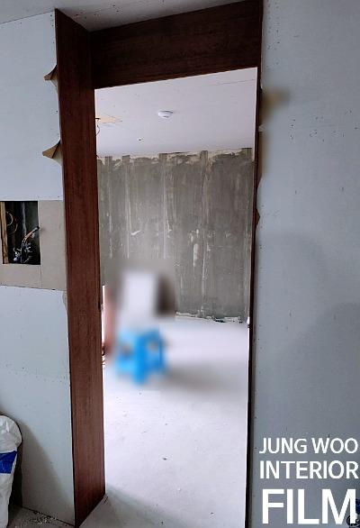 실내 공간의 벽면이 흰색 마감 처리되고, 문틀은 갈색 목재로 되어 있으며, 벽면 일부는 콘크리트 상태로 보이는데, 벽면 중앙에는 흰색 라인으로 마감된 부분이 있고, 벽면 왼쪽에는 전선이 노출된 구멍이 있으며, 바닥은