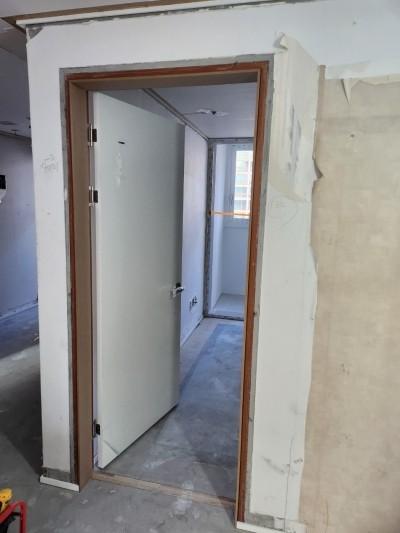 공사 중인 복도 공간, 벽면이 흰색 마감재로 덮여 있고 일부 벽면이 벗겨진 상태, 바닥은 콘크리트 원목 바닥, 문은 흰색 도어로 열려 있으며 문틀은 갈색 목재로 되어 있고 문 손잡이가 보임