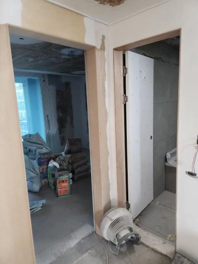 공사 중인 실내 공간으로, 벽면에 흰색 마감재가 부분적으로 붙어 있고, 문틀은 목재로 되어 있으며, 한쪽 공간에는 여러 상자와 가방이 쌓여 있고, 바닥은 콘크리트로 되어 있으며, 문 옆에는 선풍기가 놓여 있다.