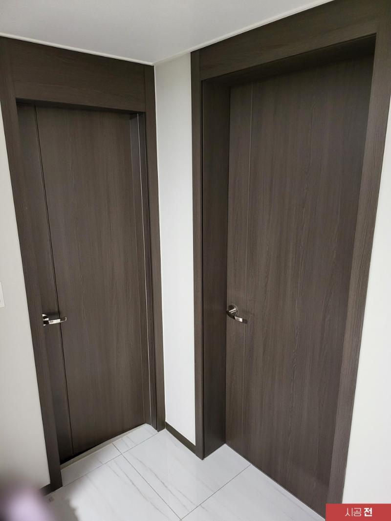 두 개의 어두운 색상의 도어가 벽에 설치된 복도 공간. 벽은 흰색, 바닥은 흰색 타일로 마감됨. 도어는 금속 손잡이가 장착된 단일 문짝 구조로, 문틀과 문짝은 어두운 목재 패턴으로 마감됨.