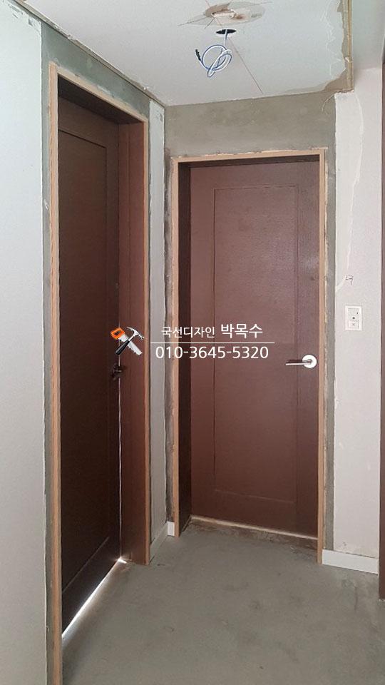 복도 공간에 두 개의 문이 설치되어 있으며, 벽면은 마감되지 않은 상태로 보입니다. 천장에는 선풍기와 전선이 노출되어 있고, 바닥은 콘크리트로 되어 있습니다. 문은 갈색으로, 문틀은 목재로 보입니다.