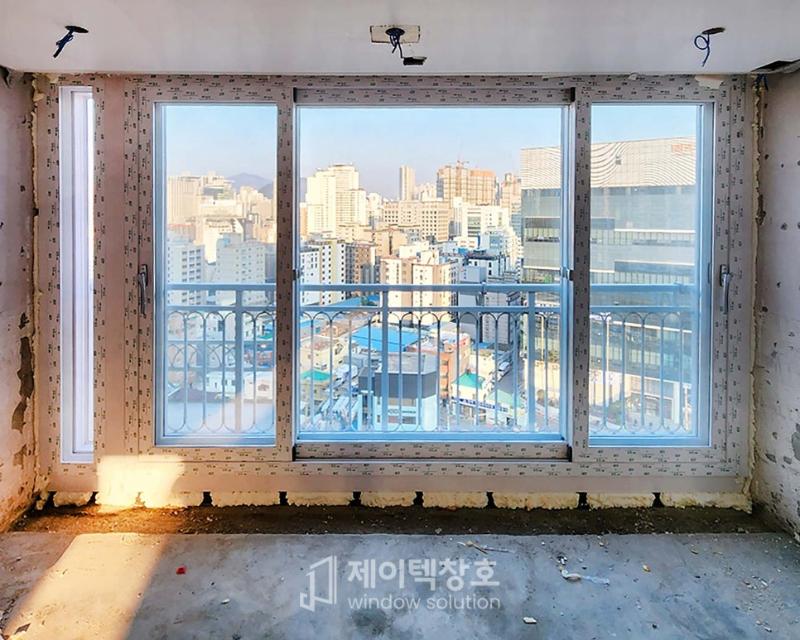 창호가 보이는 실내 공간, 벽면은 마감되지 않은 상태로 흰색 벽지가 붙지 않은 상태, 바닥은 콘크리트 바닥, 창호는 3개의 유리 패널로 구성된 슬라이딩 창호, 창호 외부에는 철제 난간이 보임
